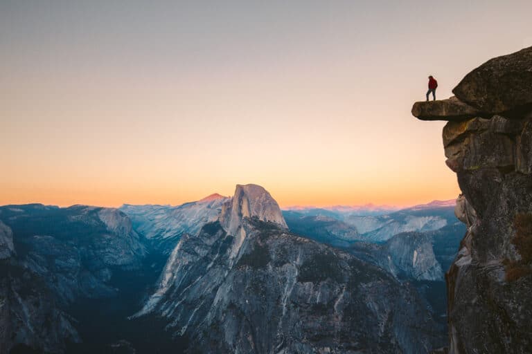 A fearless hiker is standing on an overhanging rock enjoying the view towards famous Half Dome at Glacier Point overlook in beautiful evening twilight, Yosemite National Park, California, USA