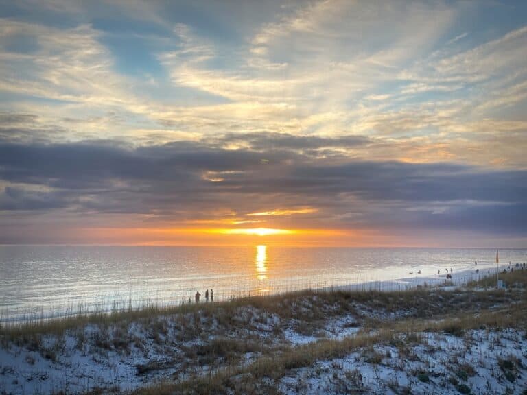 Sunset from Henderson Beach State Park, in Destin, Florida. There are a few people standing on the beach in the distance. Sea oats are growing in the white sand